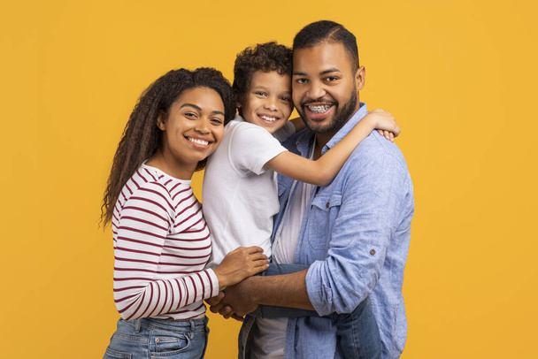 Στοργική μαύρη οικογένεια με το γιο στη μέση αγκαλιάζοντας ομαδικά, χαρούμενους νεαρούς Αφροαμερικανούς γονείς και αρσενικό παιδί ακτινοβολούν την ευτυχία, ποζάροντας μαζί σε κίτρινο φόντο στούντιο - Φωτογραφία, εικόνα