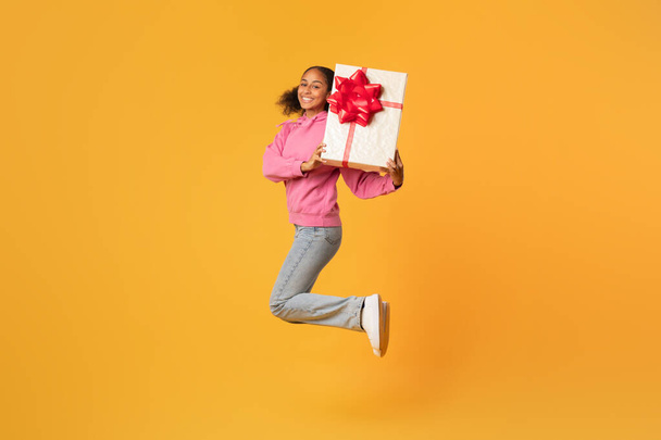 Verjaardagscadeau. Vrolijke zwarte tiener meisje springen met ingepakte geschenkdoos, glimlachen naar de camera terwijl het vieren van haar bday of speciale vakantie over gele studio achtergrond. Volledige lengte schot - Foto, afbeelding