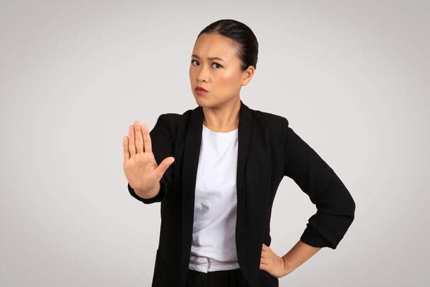 Femme d'affaires asiatique affirmée dans un blazer noir avec une expression sérieuse faisant un geste d'arrêt avec sa main, signalant frontière ou refus, sur un fond gris. Affaires et travail - Photo, image
