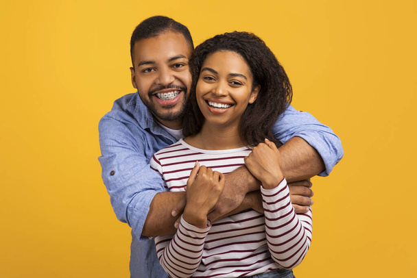 Любляча чорна пара обіймається справжніми посмішками і дивиться на камеру, щасливі молоді афроамериканські подружжя, одягнені випадково позують разом на яскраво-жовтому тлі, вільний простір - Фото, зображення