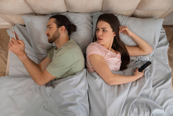 Droevige jonge vrouw wenden tot echtgenoot chatten op zijn smartphone in bed in slaapkamer interieur, bovenaanzicht. Apparaatverslaving, virtueel verraad, communicatie op afstand - Foto, afbeelding