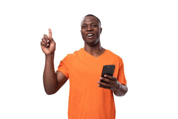 mladý Američan v oranžovém tričku drží smartphone a palce nahoru. - Fotografie, Obrázek