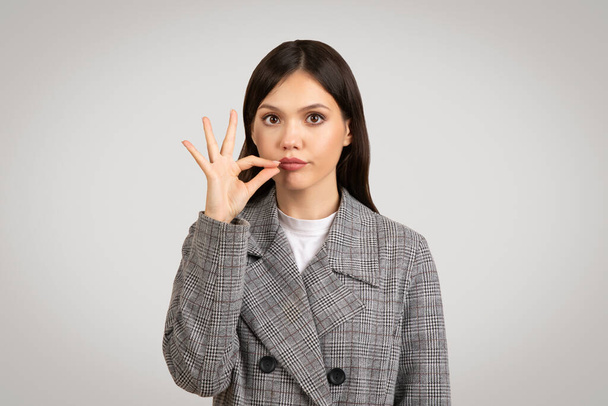 Profesionální mladá žena v šedém kostkovaném saku dělá gesto mlčení s prstem na rtech, signalizující potřebu ticha nebo utajení - Fotografie, Obrázek