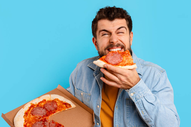 Hauska mies syö Pizza pysyvän tilalla Pizzeria laatikko sininen tausta studiossa. Nälkäinen kaveri nauttii roskaruokaa, nauttii maukasta epäterveellistä ateriaa. Huono ravitsemus tottumukset, toimitus käsite - Valokuva, kuva