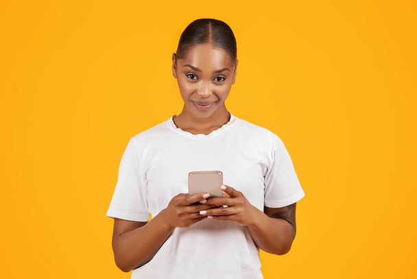モバイルアプリケーション。 黄色の背景でウェブをサーフィンするスマートフォンガジェットを使用して幸せな黒人女性,スタジオショット. ガジェットでインターネットを閲覧し,オンラインでメッセージを送信する女性 - 写真・画像