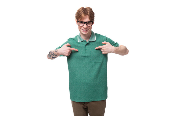 joven caucásico estudiante masculino con el pelo rojo y los puntos del tatuaje con sus manos en su camiseta verde. - Foto, Imagen