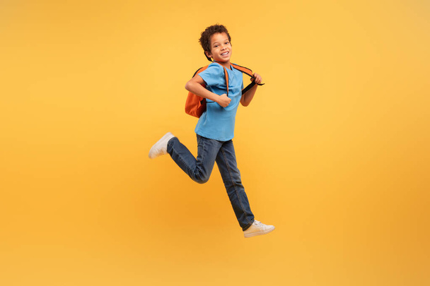 Energiczny młody chłopiec z kręconymi włosami skaczący radośnie, noszący niebieską koszulę i plecak, na dynamicznym żółtym tle, uchwycając esencję beztroskiej zabawy z dzieciństwa - Zdjęcie, obraz