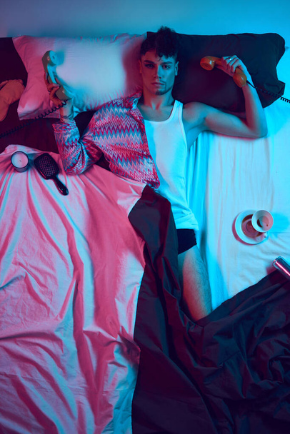 Connection. Bridging Masculine and Feminine Energies. Young male model lying in bed and holding landline retro phone and looking at camera in neon light. Concept of lifestyle, masculinity, femininity. - Photo, Image