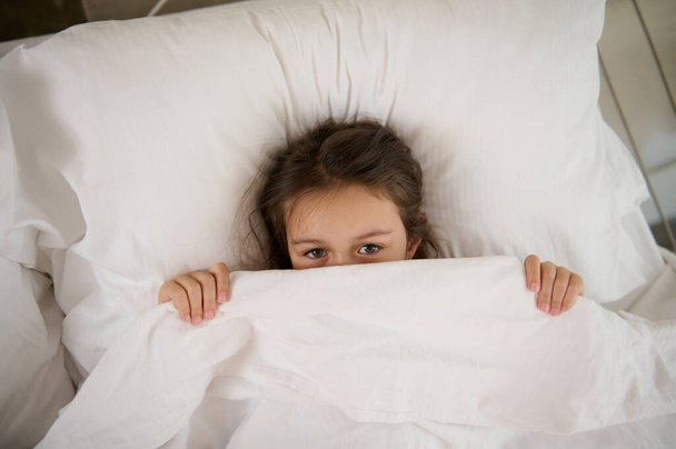 Little girl lying on the bed, hiding half of her face under a white blanket, looking at camera. Surprised child peeking from duvet wake from bad dream sleep feel fear afraid of nightmare, top view - Photo, Image