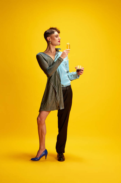 Young male model half-wearing in womans and mans clothes posing holding glasses of champagne and whiskey in hands on his female and male sides. Concept of self-expression, fashion and style, beauty. - Photo, Image