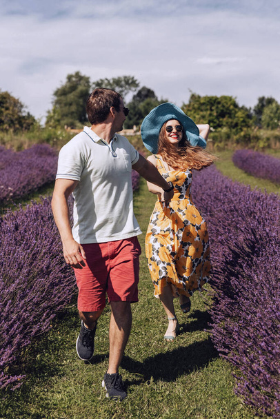 A young couple, a man and a woman, are running through a lavender field. The couple is holding hands, the man turns and looks at the woman, she smiles. A woman in a hat. A walk on the lavender meadow. - Photo, Image