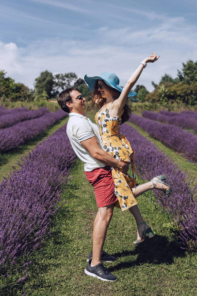 Ein verliebtes Paar, ein Mann und eine Frau, gehen auf einer Lavendelwiese spazieren. Ein Mann hält eine Frau auf dem Arm. Eine Frau mit blauem Hut lächelt ihn an und schaut ihn an. Ein Spaziergang eines jungen Paares durch ein Lavendelfeld - Foto, Bild
