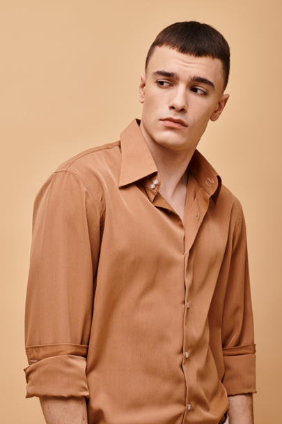Modeporträt eines stilvollen gutaussehenden Mannes im beigen Hemd, der auf pfirsichbeigem Hintergrund wegschaut - Foto, Bild