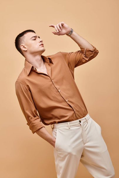 Fashion shot of stylish man in beige shirt looking away with hand in pocket on beige backdrop - Photo, Image