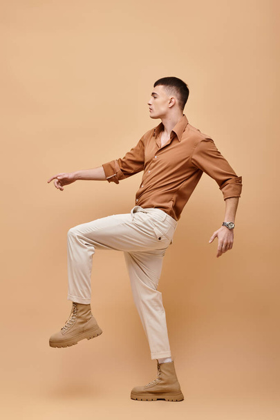 Side view photo of young man in beige shirt, pants and boots posing on beige background - Photo, Image