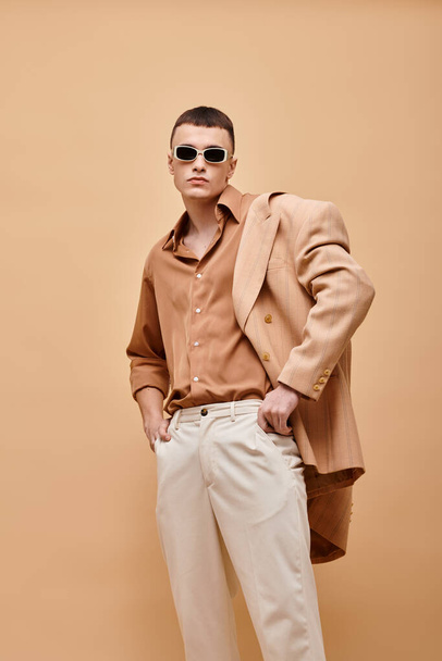 Hombre de moda en chaqueta beige en hombro, camisa, pantalones y gafas de sol posando sobre fondo beige - Foto, Imagen