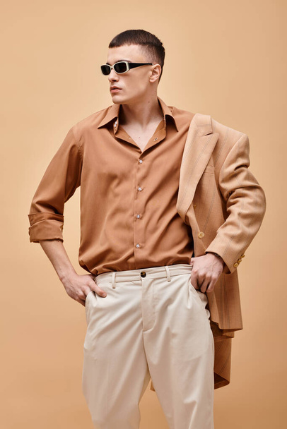 Элегантный мужчина в бежевой куртке на плече, рубашке, брюках и солнцезащитных очках позирует на бежевом фоне - Фото, изображение