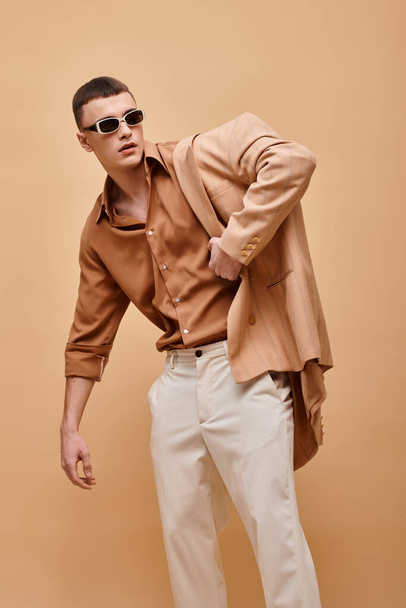 Selbstbewusster Mann in beiger Jacke auf Schulter, Hemd, Hose und Sonnenbrille posiert auf beigem Hintergrund - Foto, Bild