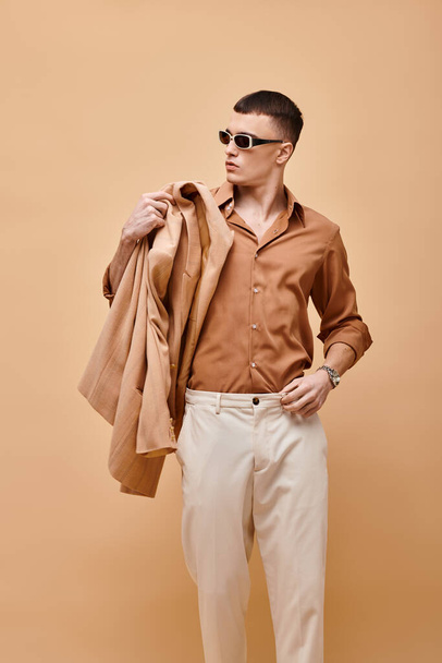 Φωτογραφία μόδας του άνδρα σε μπεζ πουκάμισο με γυαλιά ηλίου και κρατώντας σακάκι στο χέρι σε μπεζ φόντο - Φωτογραφία, εικόνα