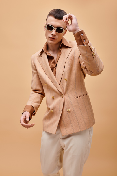 Zelfverzekerde man in beige jas, shirt, broek en zonnebril poserend op beige achtergrond - Foto, afbeelding