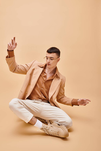 Handsome man in beige jacket and glasses sitting in lotus pose on peachy beige background - Photo, Image