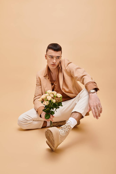 Hombre guapo en chaqueta beige y gafas sentado con rosas sobre fondo beige melocotón - Foto, Imagen