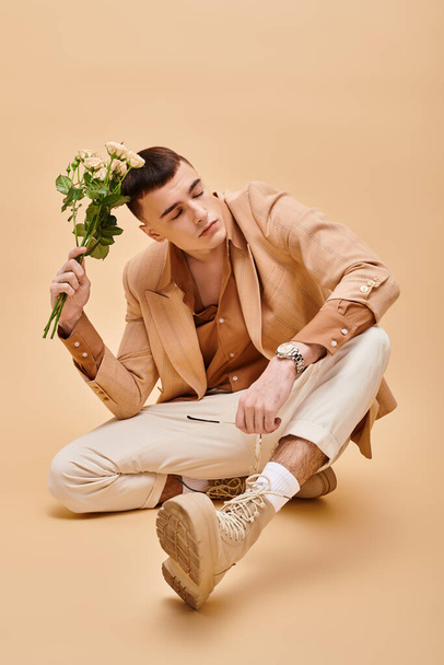 Hombre guapo en chaqueta beige sentado con rosas y gafas sobre fondo beige mirando hacia otro lado - Foto, Imagen