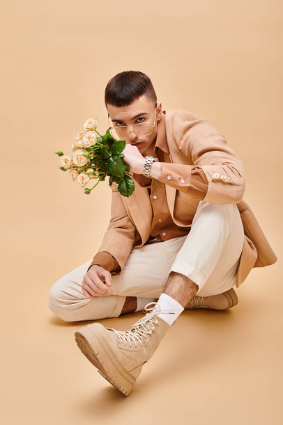 Fiatal férfi bézs dzsekiben ül rózsákkal és szemüveggel bézs háttéren a kamerába nézve - Fotó, kép
