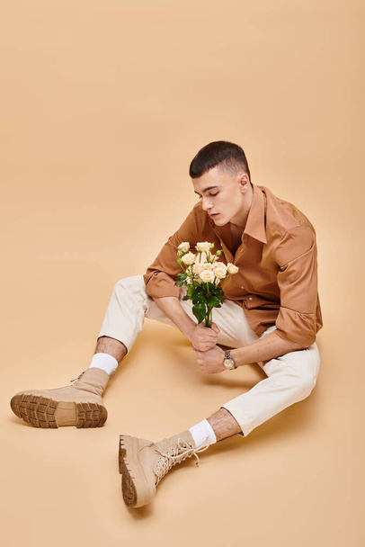 Hombre de moda en camisa beige sentado con flores y gafas sobre fondo beige mirando hacia abajo - Foto, Imagen