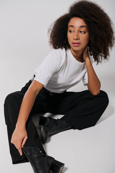 sierlijke Afrikaanse Amerikaanse vrouw in stijlvolle kleding zitten op de vloer en weg te kijken, mode concept - Foto, afbeelding