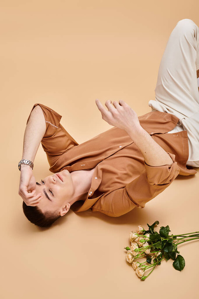 Модний портрет чоловіка в бежевій сорочці, що лежить з букетом троянд на бежевому фоні - Фото, зображення