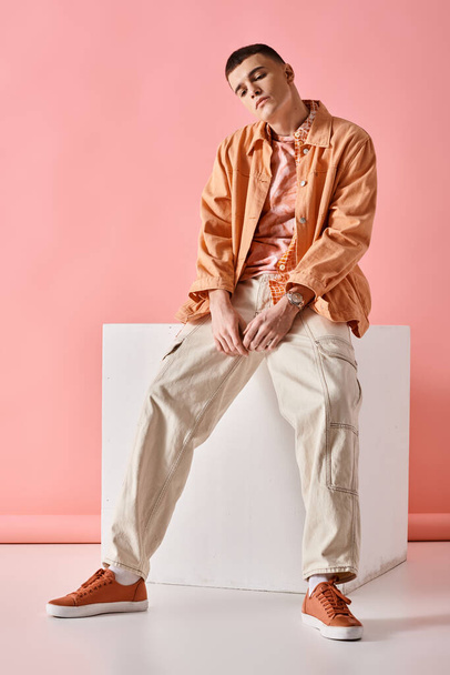 Komea mies beige paita, housut ja lenkkarit istuu valkoinen kuutio vaaleanpunainen tausta - Valokuva, kuva