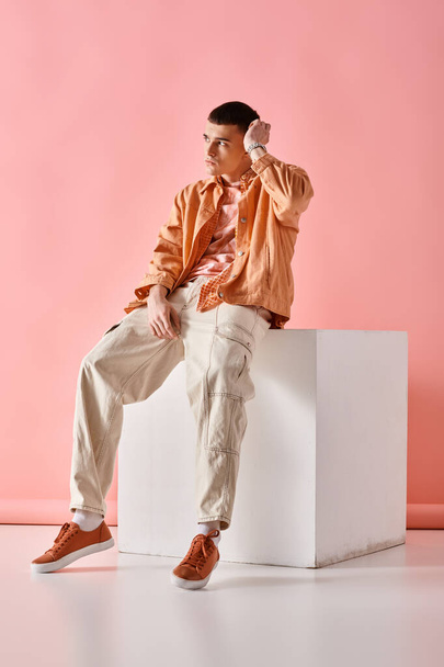 Модный мужчина в бежевой рубашке, брюках и кроссовках сидит на белом кубе на розовом фоне - Фото, изображение
