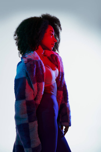 séduisante femme afro-américaine en fausse fourrure regardant loin dans les lumières rouges et bleues, concept de mode - Photo, image