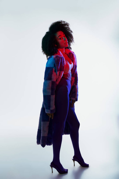 debonair african american woman in faux fur with curly hair looking at camera in red and blue lights - Photo, Image