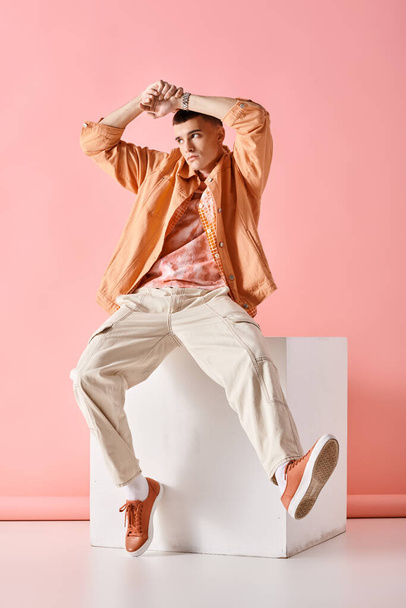 Joven de moda en traje beige tocando su cabeza y sentado en cubo blanco sobre fondo rosa - Foto, imagen