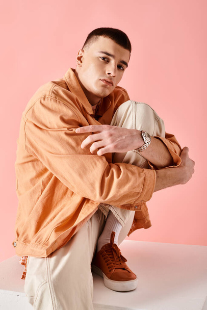 Homem de 20 anos de roupa elegante abraçando o joelho escondido e sentado no cubo branco no fundo rosa - Foto, Imagem