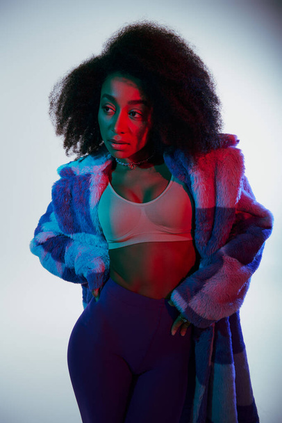 sexy chic donna africana americana in ecopelliccia vibrante e reggiseno guardando lontano in luci rosse e blu - Foto, immagini