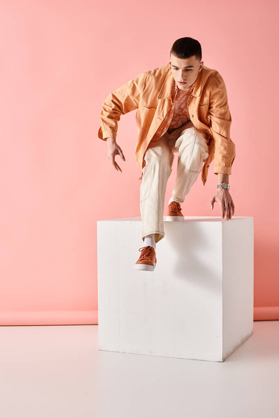 Porträt eines stilvollen Mannes im beigen trendigen Outfit, der von einem weißen Würfel auf rosa Hintergrund springt - Foto, Bild