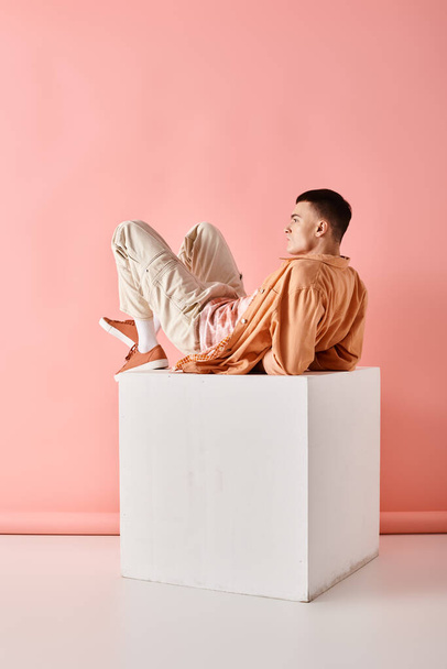 Πλάγια φωτογραφία του άνδρα σε ροδακινί χρώμα στολή που βρίσκεται σε λευκό κύβο σε ροζ φόντο - Φωτογραφία, εικόνα