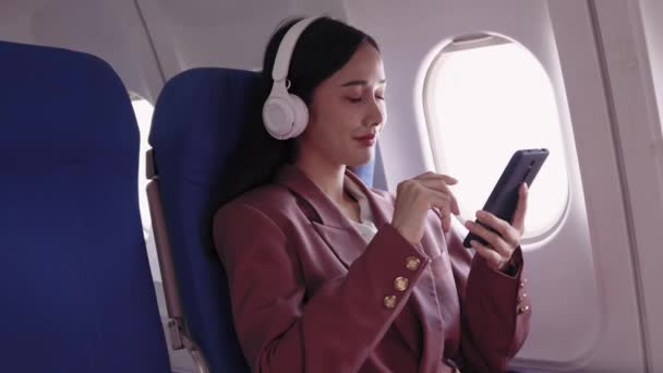Aziatisch zakenvrouw kiest liedjes op haar mobiele telefoon en geniet van haar favoriete muziek via over-ear hoofdtelefoon tijdens het reizen op een vliegtuig.  - Video
