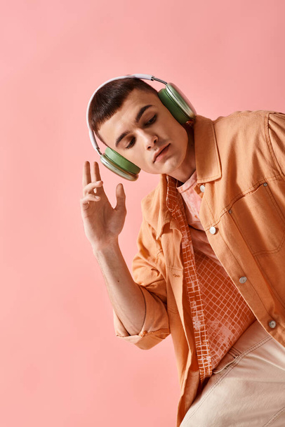 Modny mężczyzna w stroju warstwowym ze słuchawkami bezprzewodowymi słuchający muzyki na różowym tle - Zdjęcie, obraz