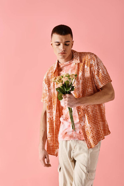 Fashion shot of fashionable man in layered outfit holding flowers on pink backdrop - Photo, Image