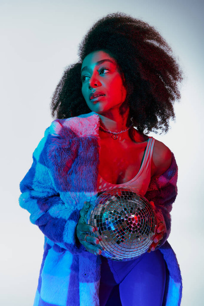 séduisante femme afro-américaine élégante en fausse fourrure posant avec boule disco et détournant les yeux - Photo, image