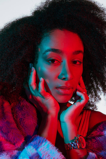 attraente donna afro-americana alla moda in eco-pelliccia guardando la fotocamera a luci rosse e blu - Foto, immagini