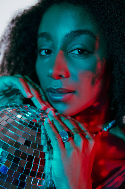 séduisante femme afro-américaine à la mode en fausse fourrure regardant la caméra en lumières rouges et bleues - Photo, image