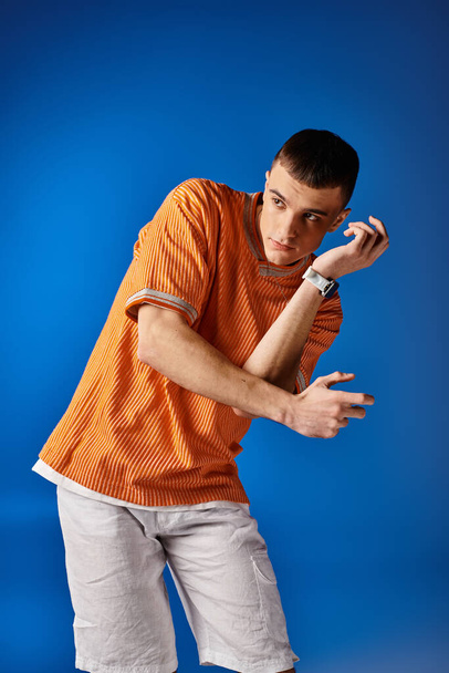 Модний знімок стильного чоловіка в помаранчевій сорочці та білих шортах, що рухаються на синьому фоні - Фото, зображення