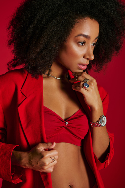 σέξι αφροαμερικανή γυναίκα σε κόκκινο κομψό ντύσιμο κοιτάζοντας μακριά στο κόκκινο φόντο, έννοια της μόδας - Φωτογραφία, εικόνα