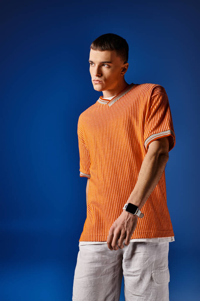 Stijlvolle jonge man in trendy oranje shirt en witte shorts die wegkijken op diepblauwe achtergrond - Foto, afbeelding