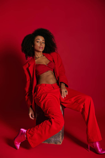 atraente mulher americana africana sexy em traje vermelho sentado e olhando para a câmera, conceito de moda - Foto, Imagem
